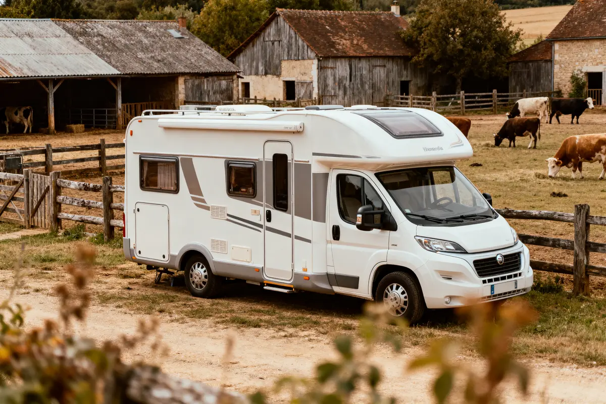 Camping-car blanc stationné dans une ferme, avec grange, vaches broutant, et clôture en bois environnante.