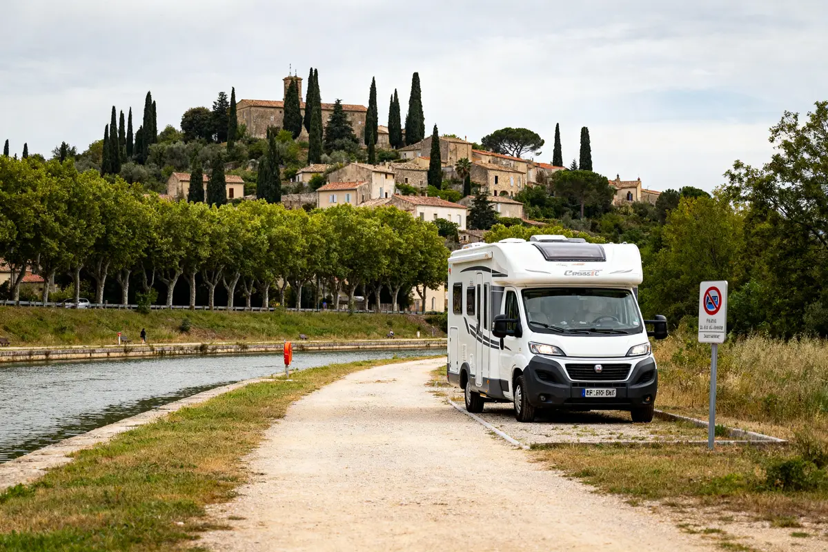 Camping-car stationné près d'un canal, village provençal en arrière-plan avec cyprès et bâtiments historiques.