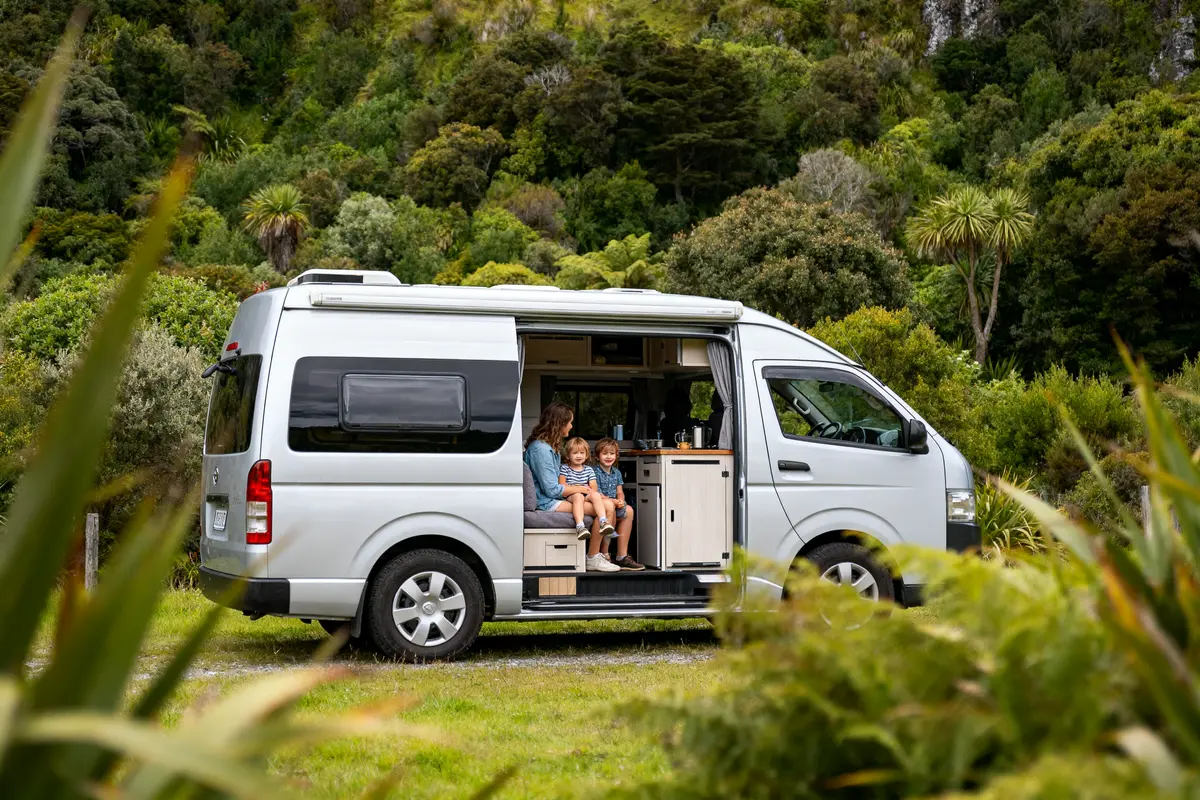 Camping-car blanc, famille assise à l'intérieur, forêt verdoyante en arrière-plan, journée ensoleillée.