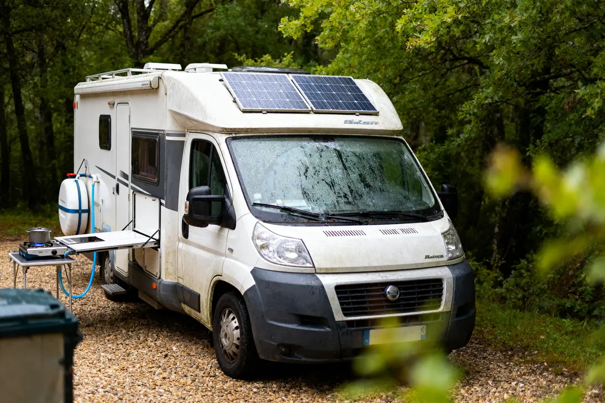 Camping-car blanc avec panneaux solaires stationné en forêt, table et réchaud à gaz à l'extérieur.