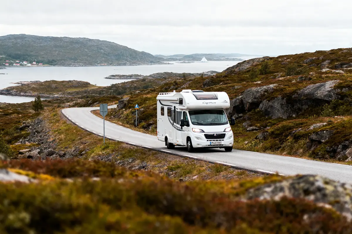 Camping-car blanc traversant une route sinueuse parmi des collines verdoyantes, avec un lac en arrière-plan.