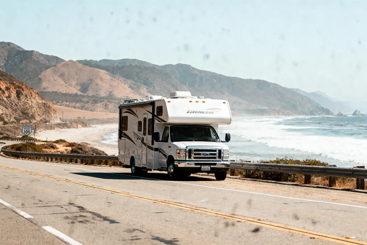 Camping-car roulant sur une route côtière, falaise et océan en arrière-plan, journée ensoleillée.