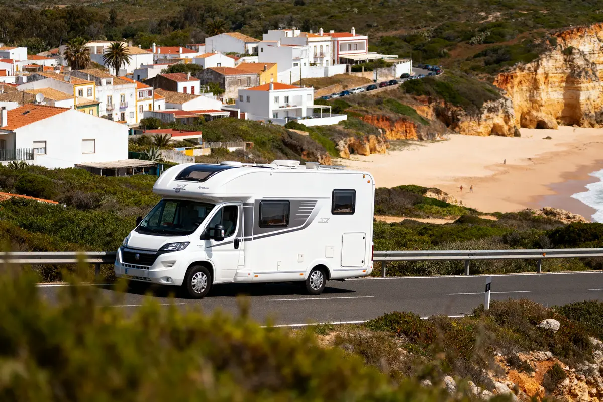 Camping-car roulant sur route, village en arrière-plan, plage et falaises sous un ciel clair.