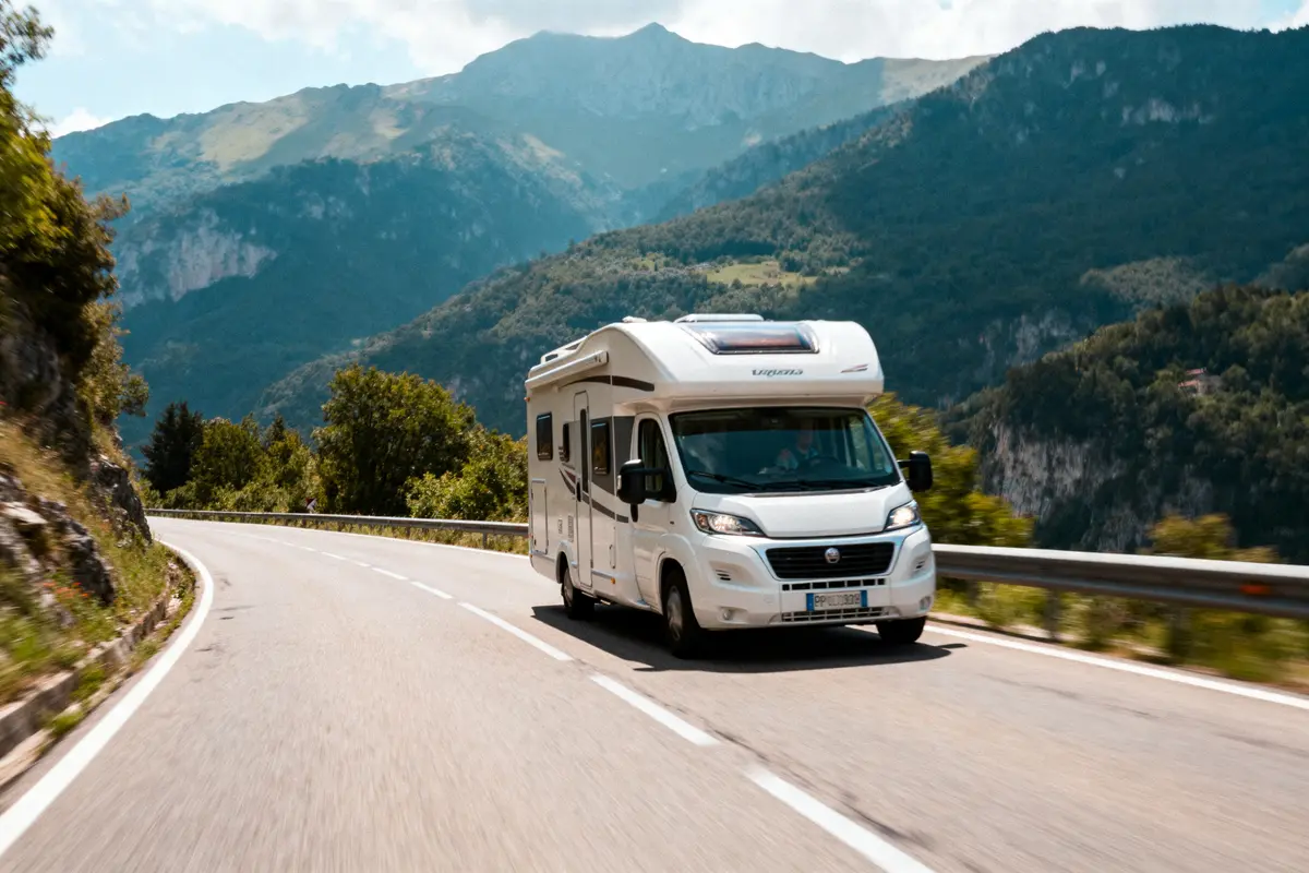 Camping-car blanc roulant sur une route montagneuse, entouré de paysages verdoyants et montagnes en arrière-plan.