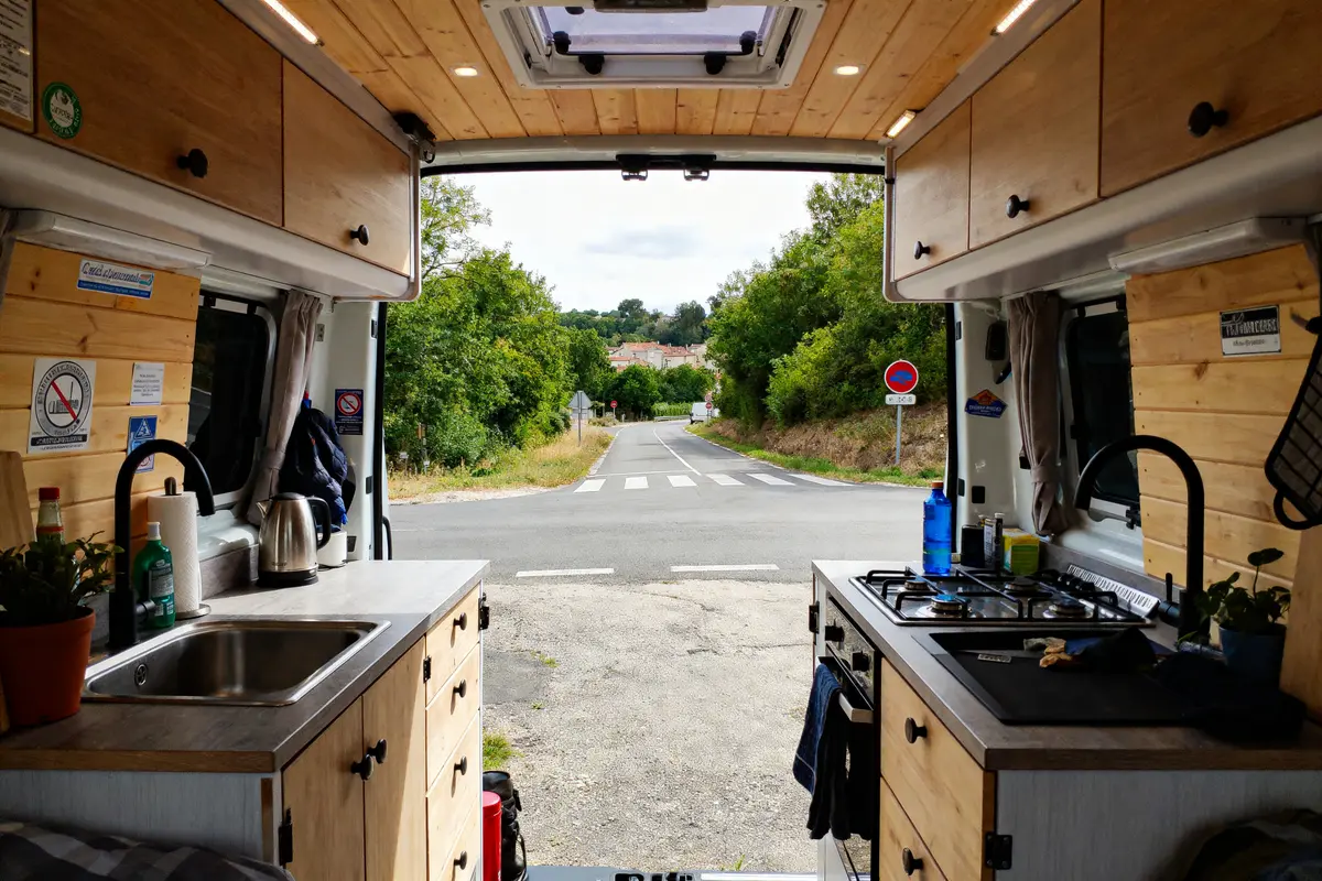 Intérieur de van aménagé avec cuisine, vue route et végétation à l'extérieur.