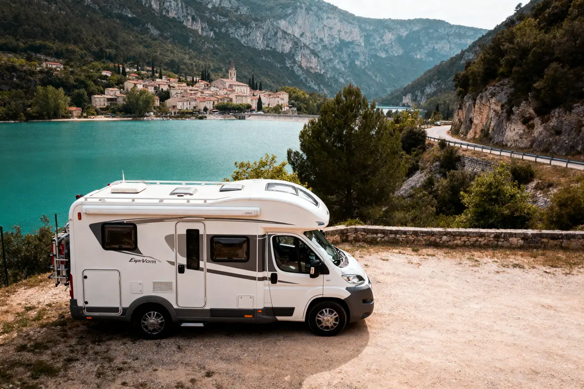 Camping-car garé près d'un lac turquoise, entouré de montagnes et village pittoresque en arrière-plan.