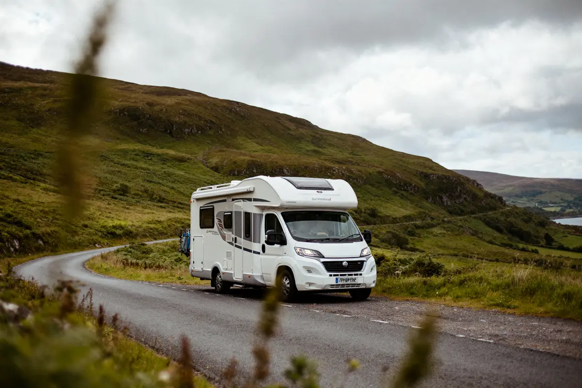 Camping-car blanc sur route sinueuse, entouré de collines verdoyantes sous un ciel nuageux. Paysage rural.