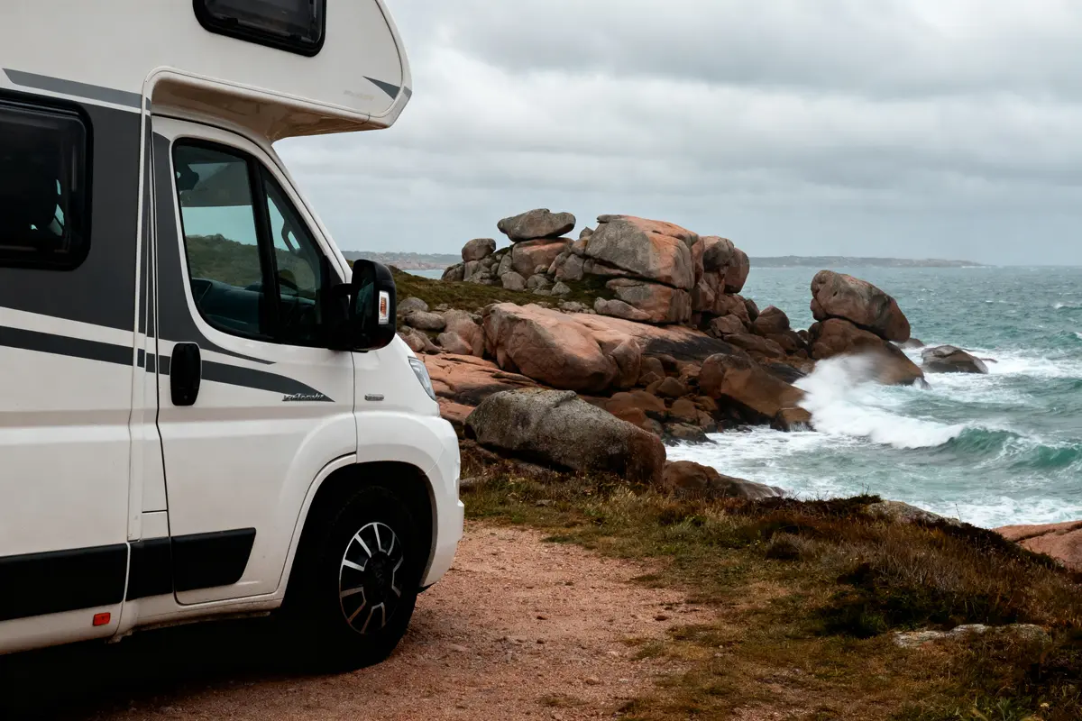 Camping-car blanc en bord de mer, rochers roses, vagues s'écrasant, ciel nuageux.