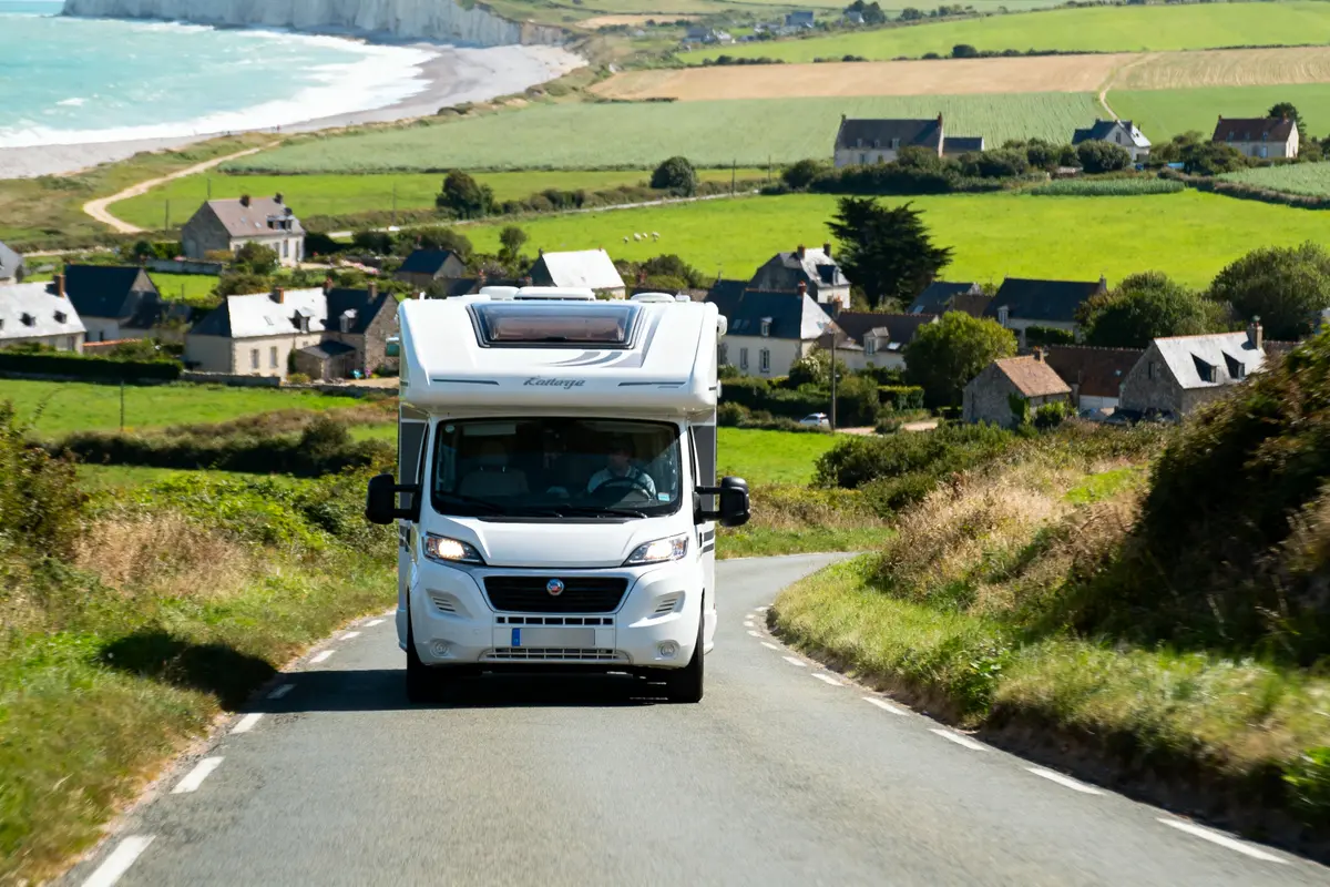Camping-car blanc sur route de campagne avec village et mer en arrière-plan. Paysage verdoyant et colline.