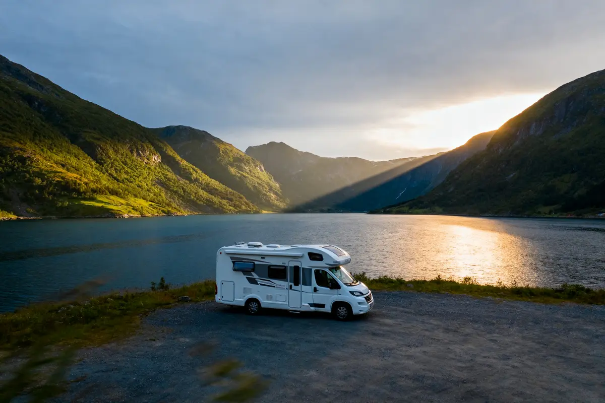 Camping-car près d'un lac entouré de montagnes, soleil couchant illuminant le paysage pittoresque.