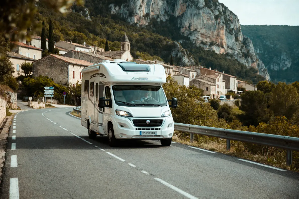 Camping-car blanc sur route sinueuse près d'un village pittoresque, entouré de collines verdoyantes et arbres.