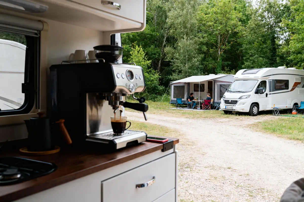 Vue intérieure d'un camping-car avec machine à café, extérieur avec camping-car, auvent et table de camping.