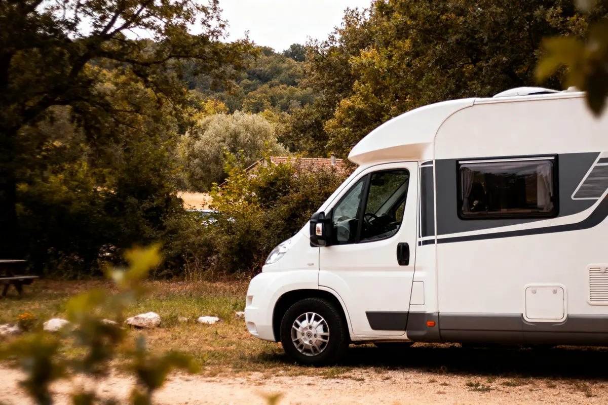 Camping-car blanc stationné dans une clairière, entouré d'arbres et verdure dense, par une journée ensoleillée.