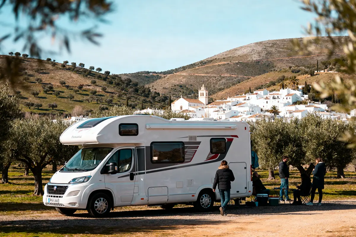 Camping-car stationné près d'un village blanc, gens réunis, collines et oliviers en arrière-plan.