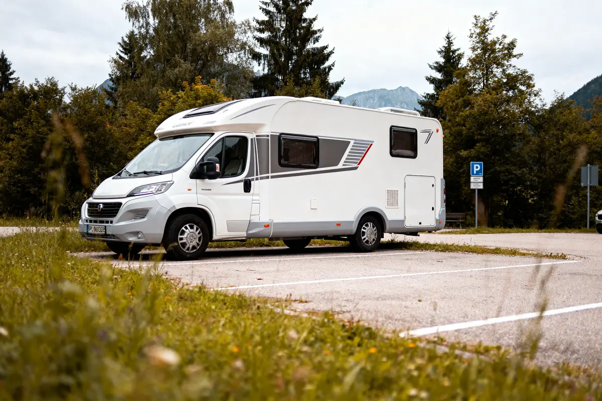 Camping-car blanc garé sur un parking entouré de verdure et d'arbres, avec montagnes en arrière-plan.