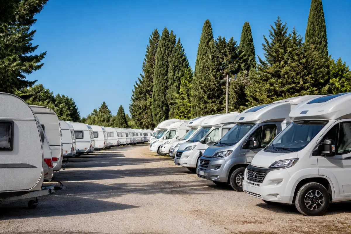 Alignement de caravanes et camping-cars sur un terrain, entouré d'arbres et ciel bleu.