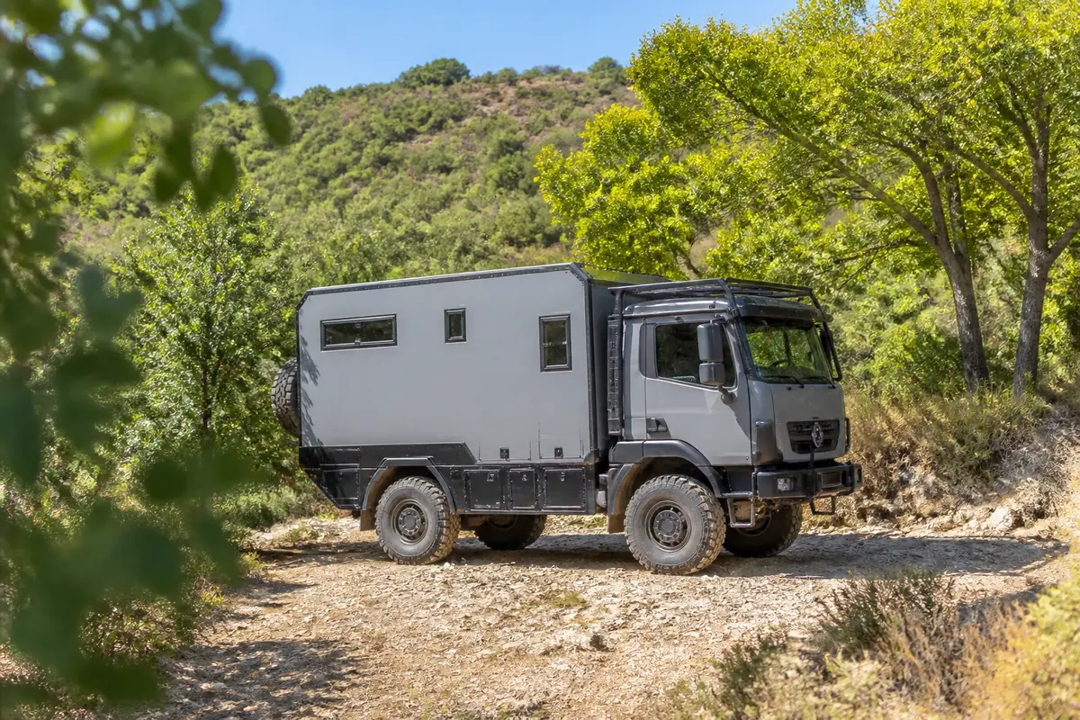 Camion d'expédition sur chemin de terre, entouré de verdure et montagnes en arrière-plan. Journée ensoleillée.