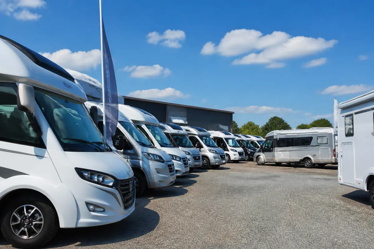 Rangée de camping-cars garés sur un parking, ciel bleu et nuages blancs en arrière-plan.