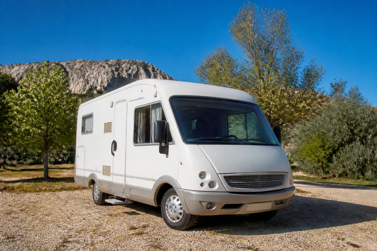 Camping-car blanc stationné en plein air, ciel bleu, avec montagne et arbres en arrière-plan.