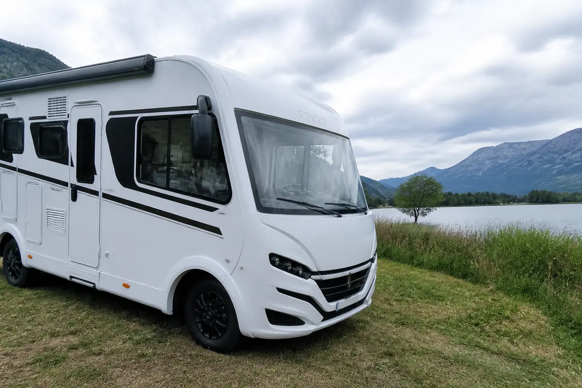 Camping-car blanc stationné près d'un lac, montagnes en arrière-plan sous ciel nuageux.
