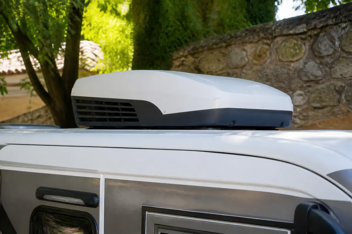 Unité de climatisation blanche installée sur le toit d'un camping-car garé, avec arbres et mur en arrière-plan.