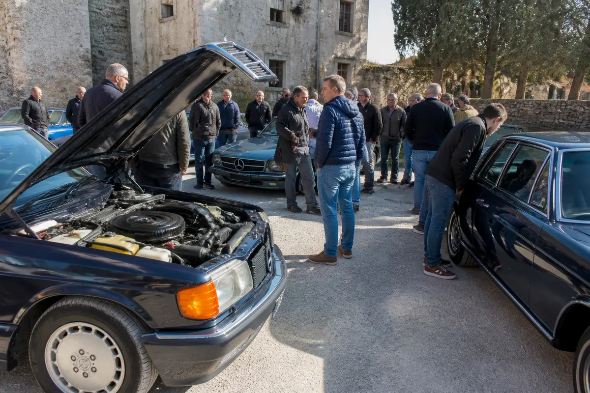 Un groupe de personnes observe des voitures anciennes avec moteur visible, discussion en extérieur ensoleillé.