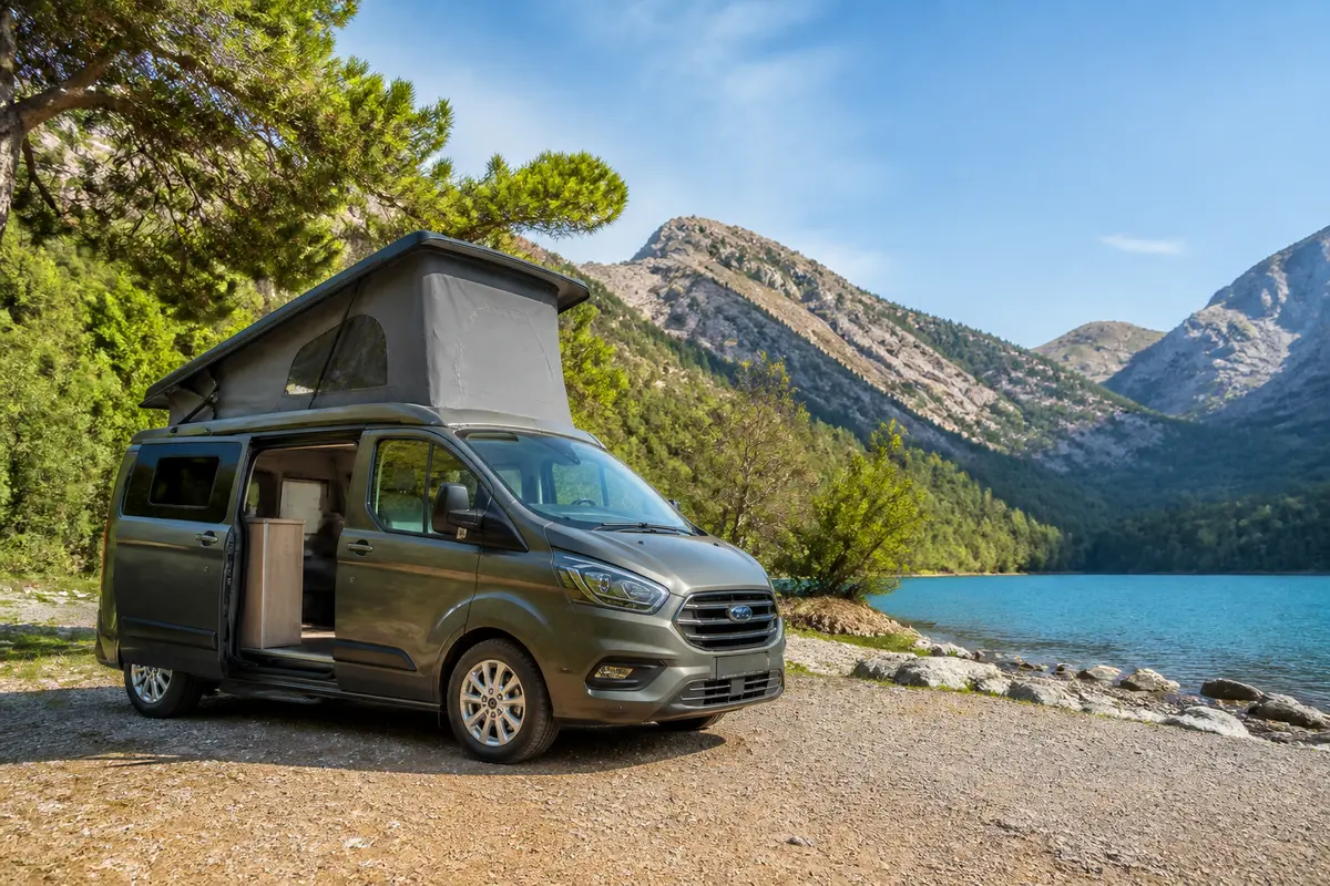 Camping-car avec toit relevé stationné près d'un lac, entouré de montagnes et d'arbres, ciel dégagé.