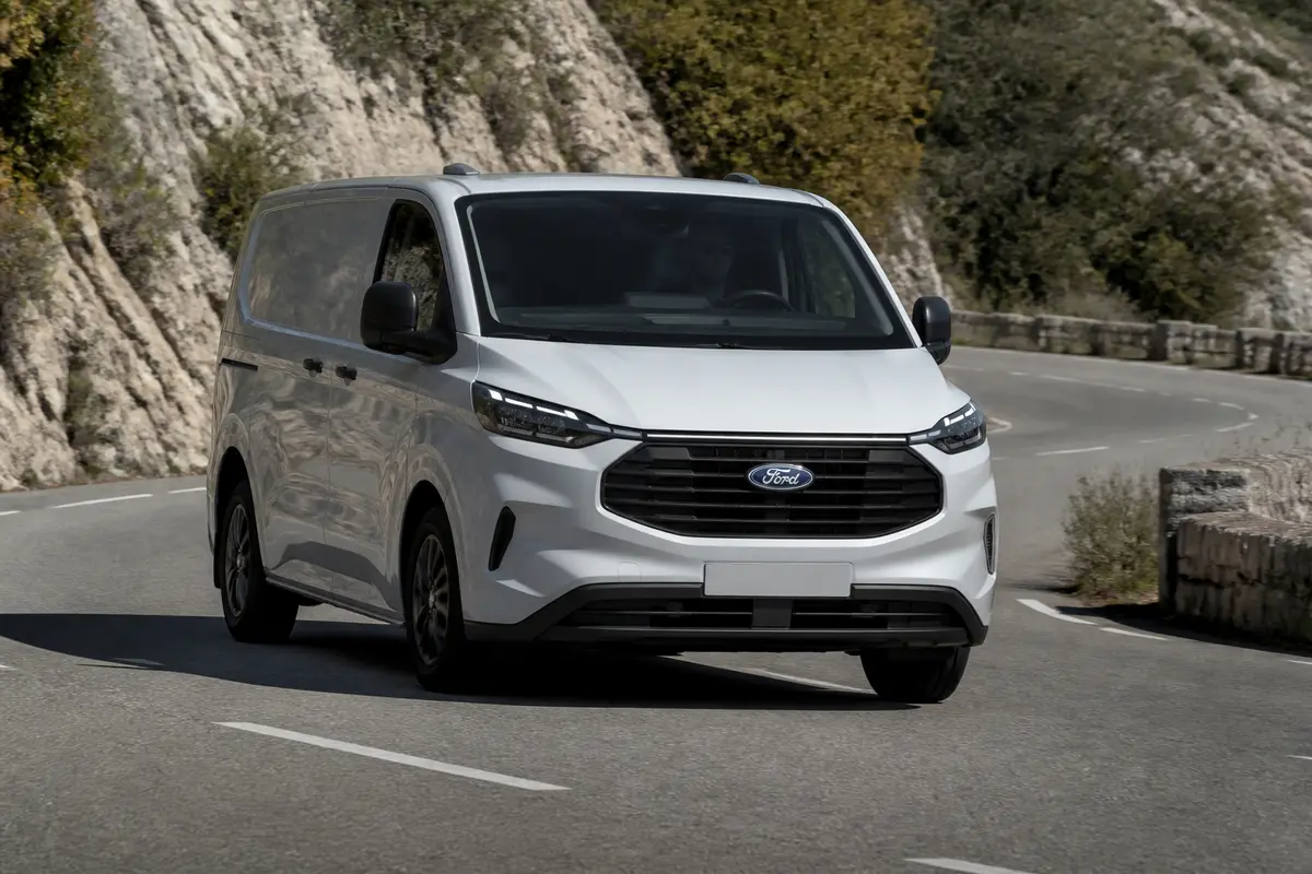 Fourgonnette Ford blanche roulant sur une route montagneuse bordée de falaises et de végétation.