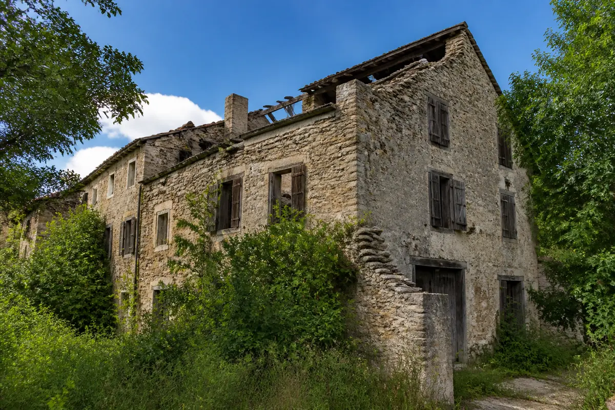 Vieille maison en pierre abandonnée, entourée de végétation, sous un ciel bleu clair.